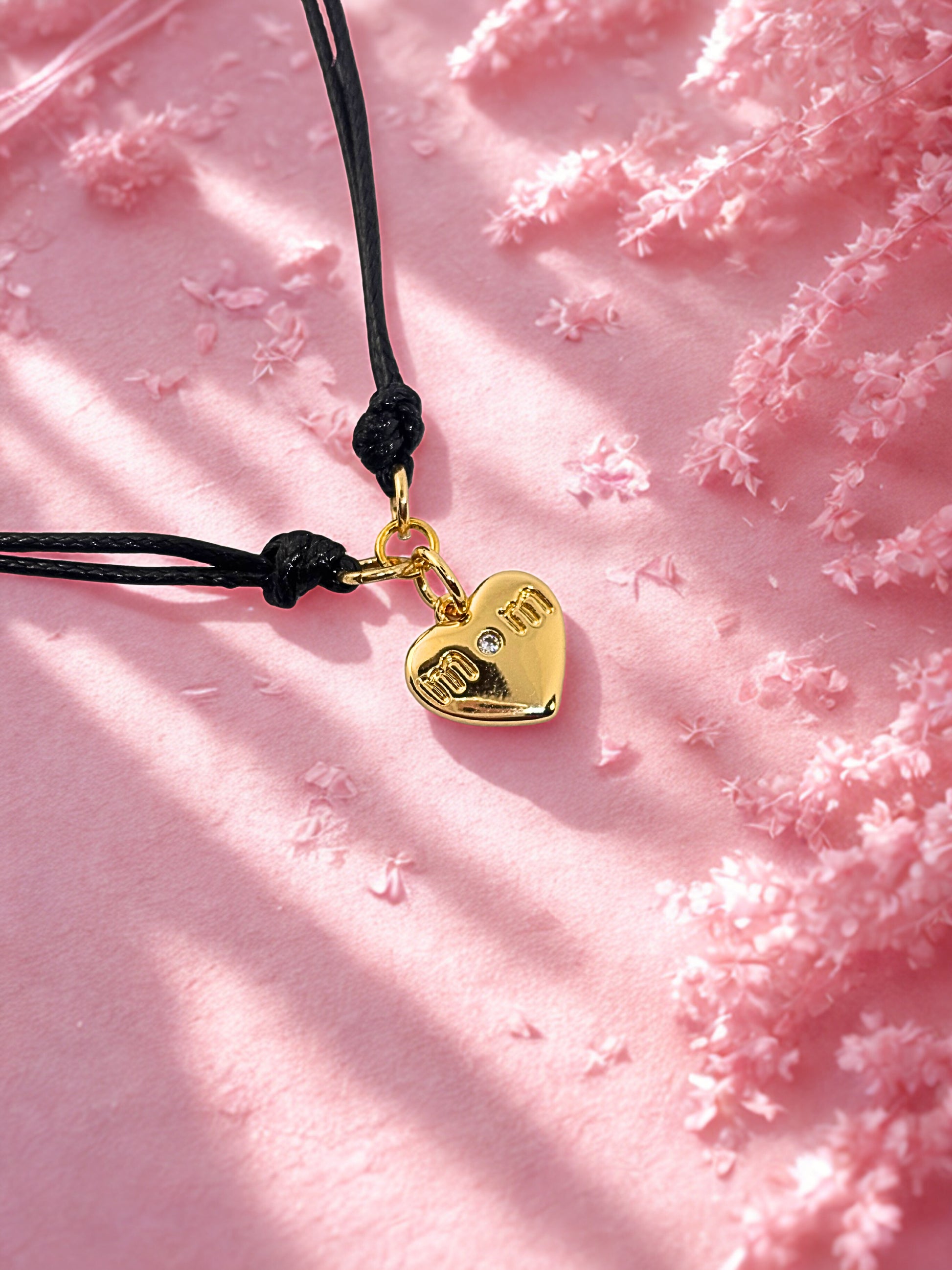 Gold heart-shaped charm bracelet on a black cord against a pink floral background
