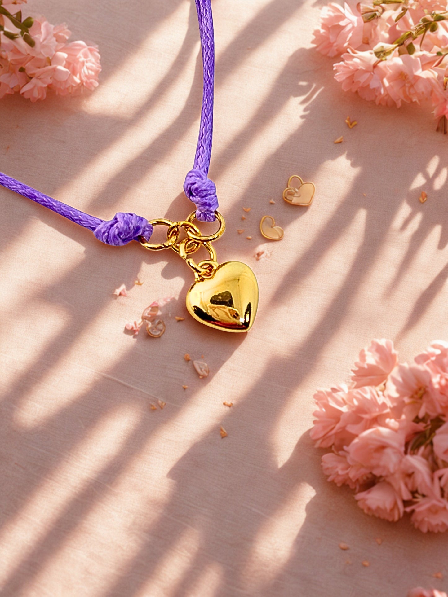 Gold heart-shaped charm bracelet on a purple cord with pink flowers in the background