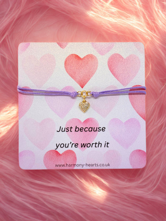 Bracelet with a heart charm on a card with heart designs and text, against a pink fluffy background.