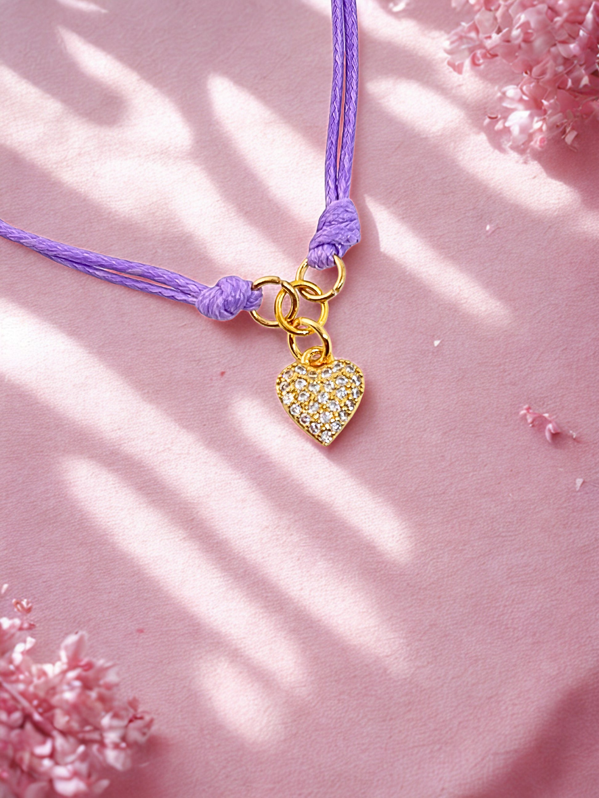 Purple cord bracelet with a gold heart charm on a pink background