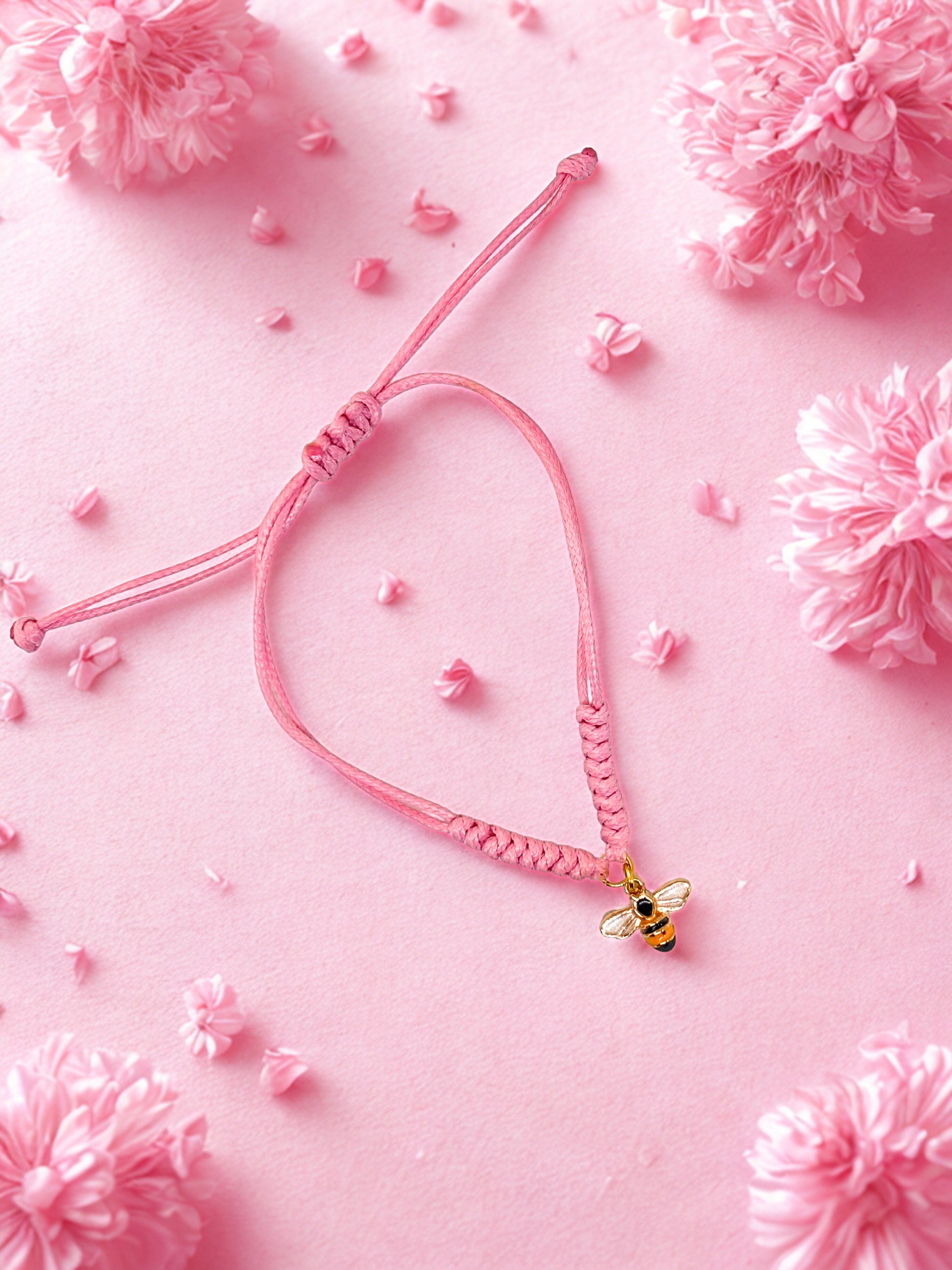 Pink braided bracelet with a bee charm on a pink background with decorative flowers.