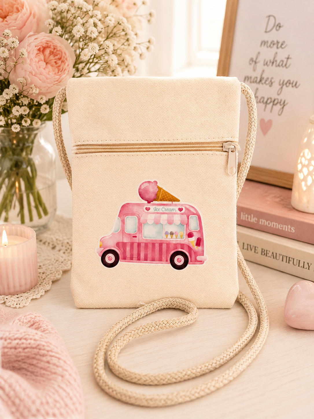 A cream-colored canvas crossbody bag featuring a pink ice cream truck illustration on the front, styled on a light wooden table with pink roses, candles, and books in the background.