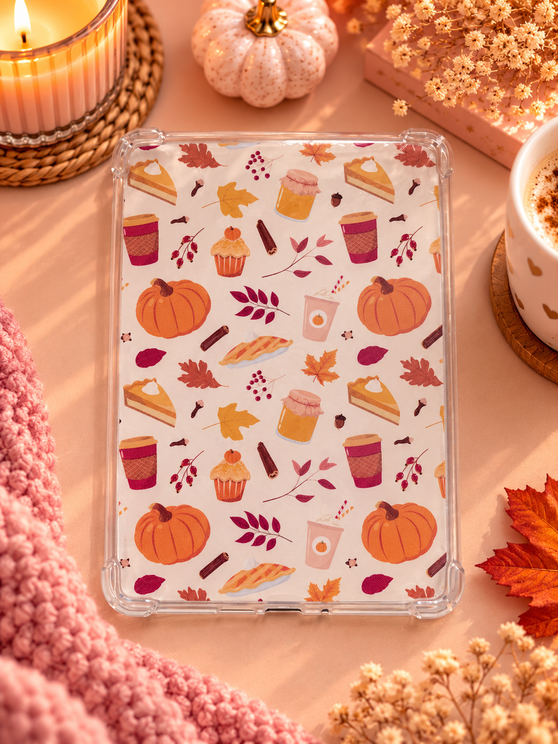 A clear kindle case with an autumn-themed print of pumpkins, leaves, cupcakes, and coffee cups, displayed in a cozy flat-lay setting with a candle, knit blanket, and dried flowers.