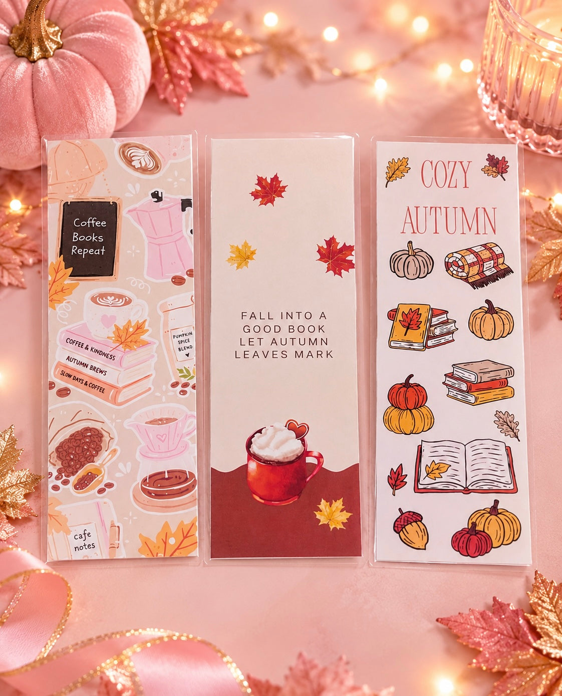 Three autumn-themed bookmarks displayed on a pink surface with warm string lights and fall decorations. The designs include illustrations of books, coffee cups, pumpkins, acorns, and leaves, with text reading 'Coffee Books Repeat,' 'Fall Into a Good Book Let Autumn Leaves Mark,' and 'Cozy Autumn.