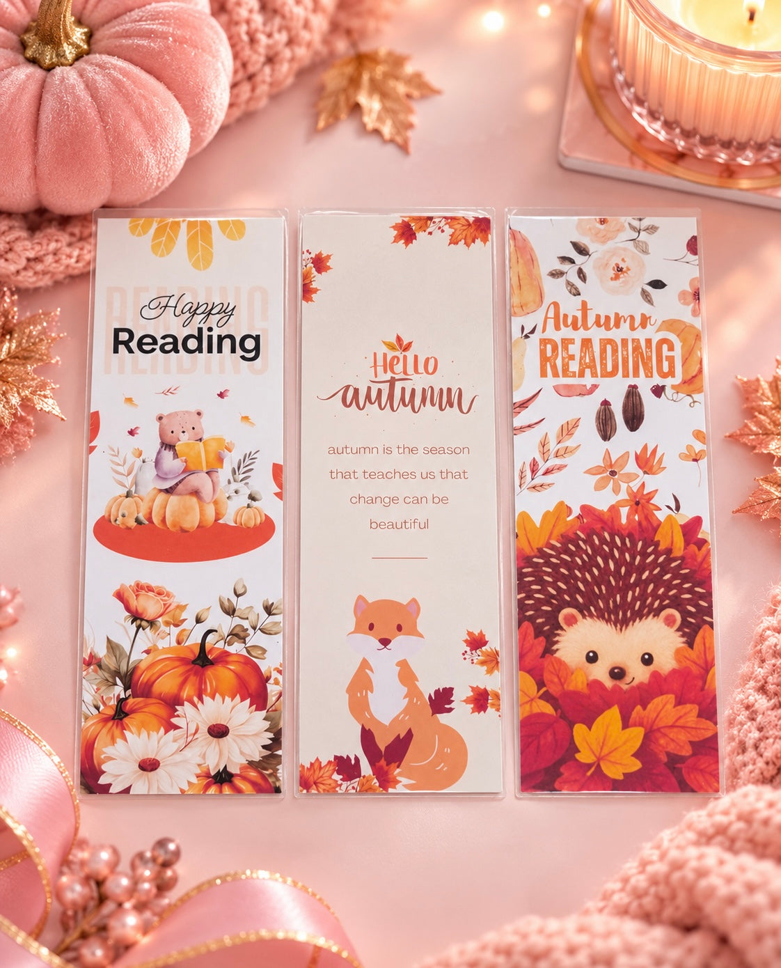 Three decorative bookmarks with autumn-themed illustrations are arranged on a pink surface with pumpkins, ribbons, and a candle. The bookmarks read 'Happy Reading,' 'Hello autumn,' and 'Autumn Reading,' and feature fall leaves, pumpkins, flowers, a fox, a hedgehog, and cozy reading imagery.