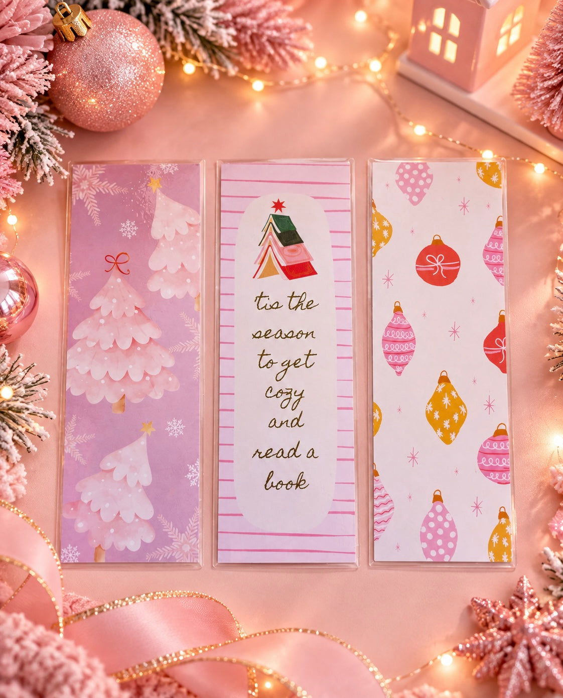 Three Christmas-themed bookmarks are displayed on a pink holiday background decorated with lights, ornaments, and ribbon. The left bookmark features a pink winter tree design, the middle bookmark has a small tree illustration and the phrase 'tis the season to get cozy and read a book', and the right bookmark shows pink and gold ornaments on a white background.
