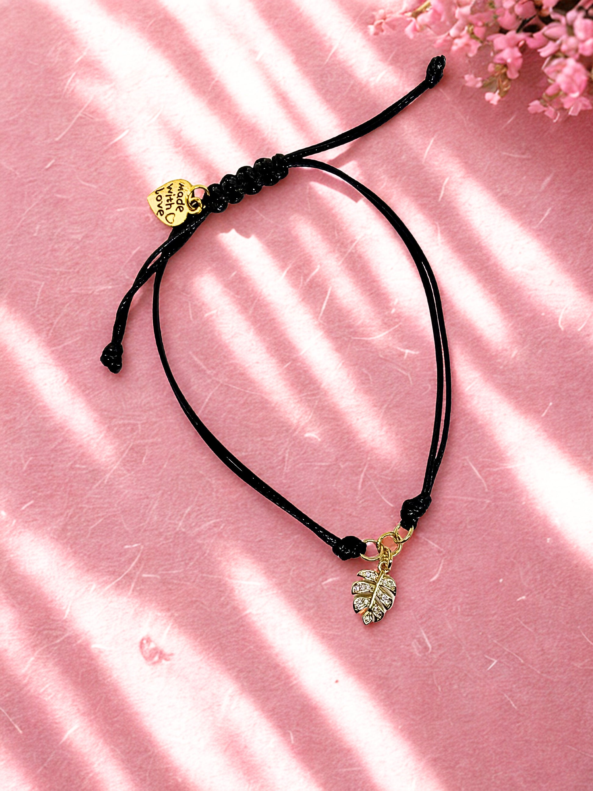 Black cord bracelet with gold leaf charm on a pink surface