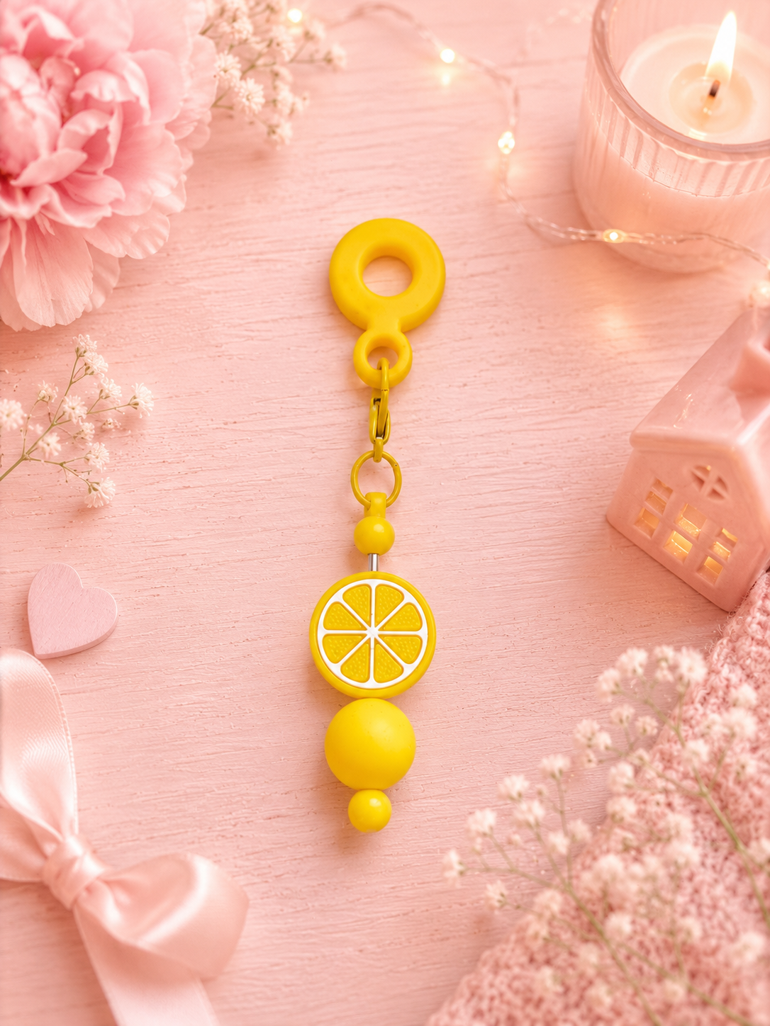 A yellow silicone cup charm with a lemon slice design and bead accents, displayed on a pink wooden surface surrounded by decorative flowers, a candle, and a small pink house figurine.
