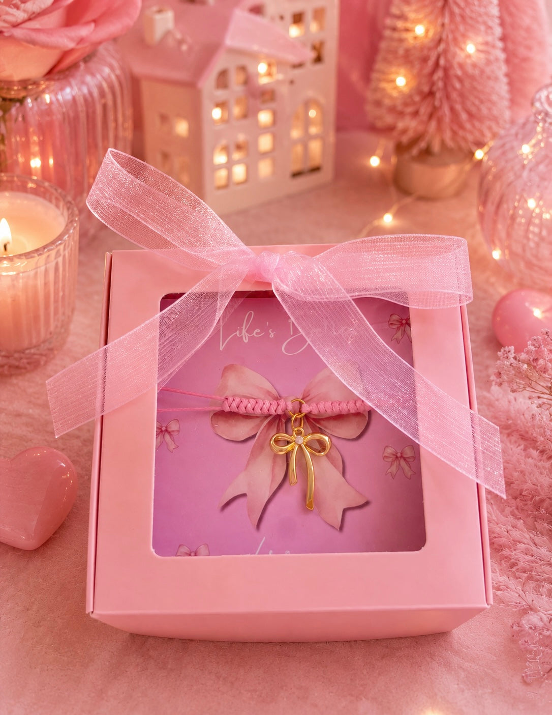 A pink gift box with a clear window, tied with a sheer pink ribbon, containing a pink braided bracelet with a small gold bow charm. The scene is styled in soft pink tones with candles, fairy lights, and decorative ornaments.