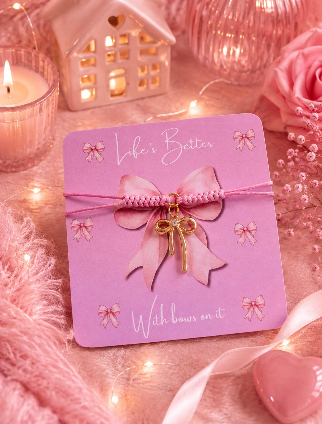 A pink carded charm bracelet with a pink braided cord and a gold bow charm, displayed on a pink background with decorative candles, fairy lights, and pink roses.
