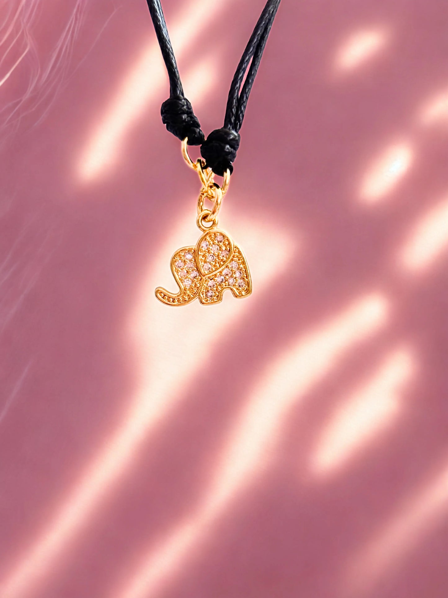Gold elephant charm on a black cord against a pink background