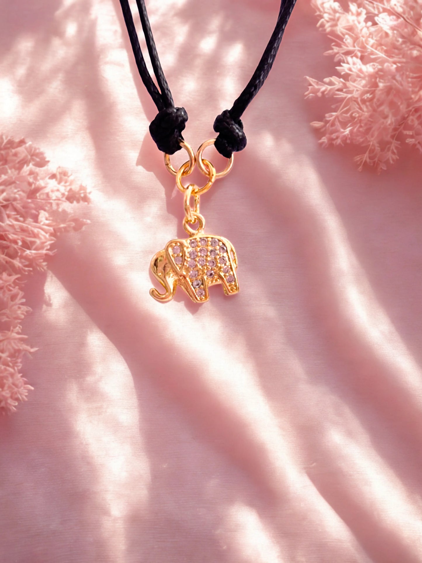 Gold elephant charm on a black cord against a pink textured background