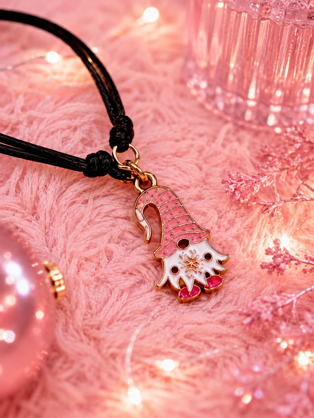 A close-up of a black cord bracelet with a gnome-shaped charm. The charm features a pink and white textured hat, a white face with gold details, and small pink shoes, resting on a pink fuzzy surface with warm fairy lights and decorative elements in the background.