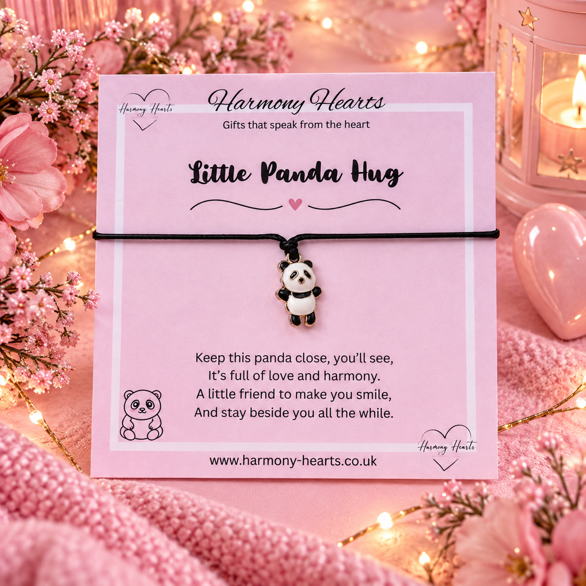 A charm bracelet with a small panda pendant on a black cord, displayed on a pink card from Harmony Hearts. The card is styled with pink flowers, fairy lights, and a heart-shaped candle holder in a cozy setting.