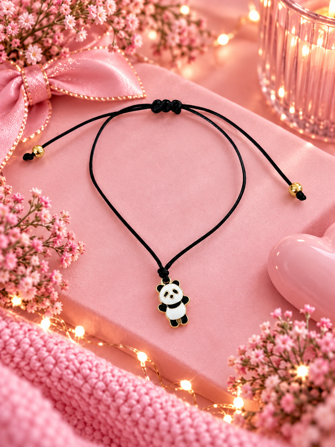 A black cord bracelet with a small panda charm, styled on a pink gift box with pink flowers, a pink ribbon, and warm fairy lights.