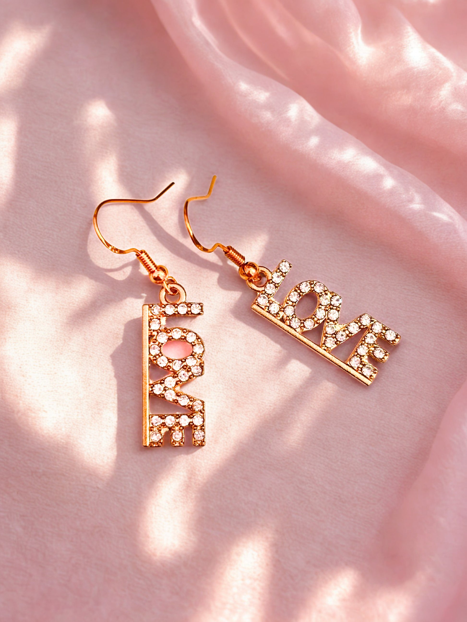 Gold sparkly earrings with the word ‘Love’ on a pink background 