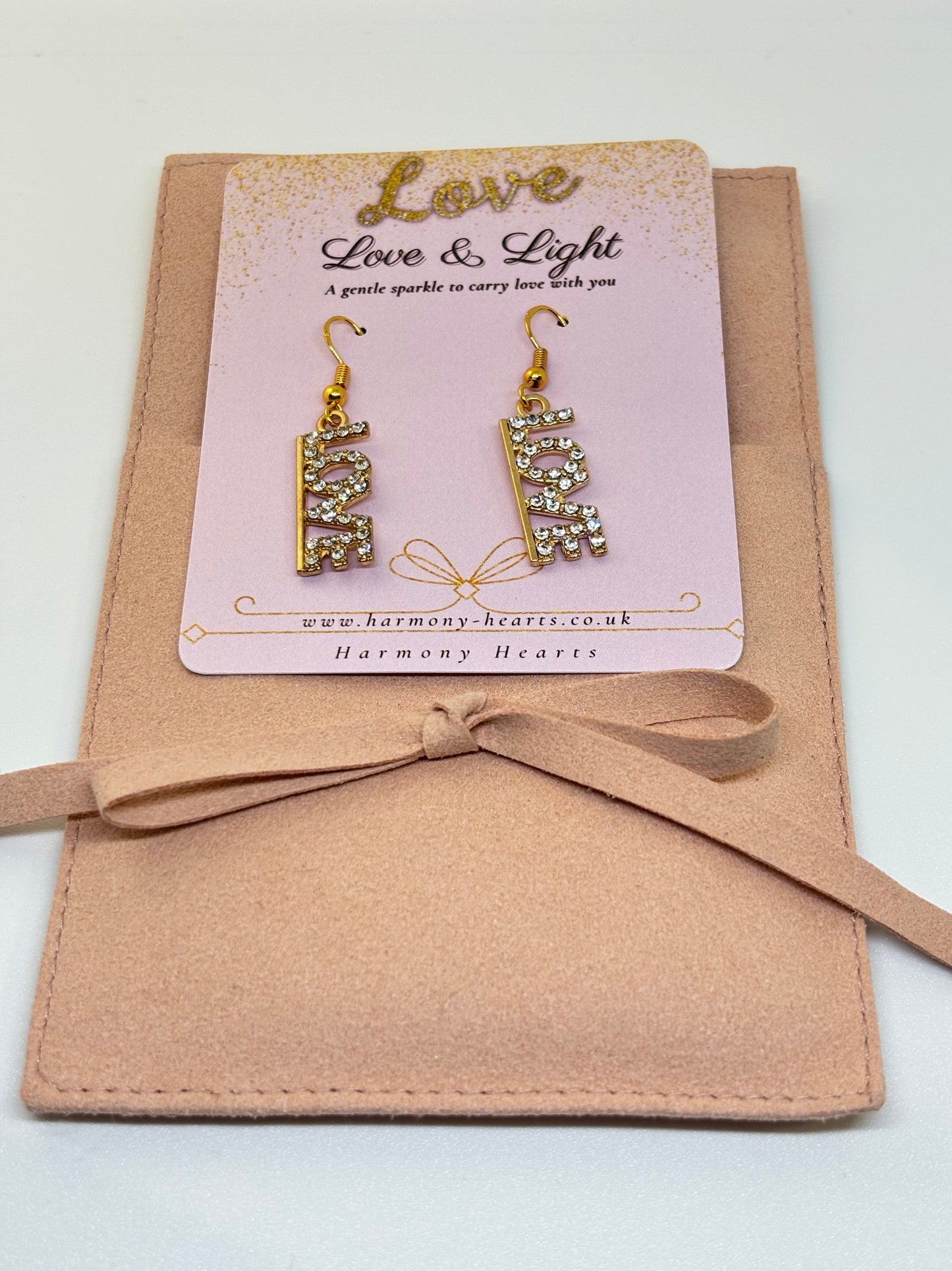 Gold earrings on a 'Love & Light' card with a pink background, placed on a beige fabric surface.