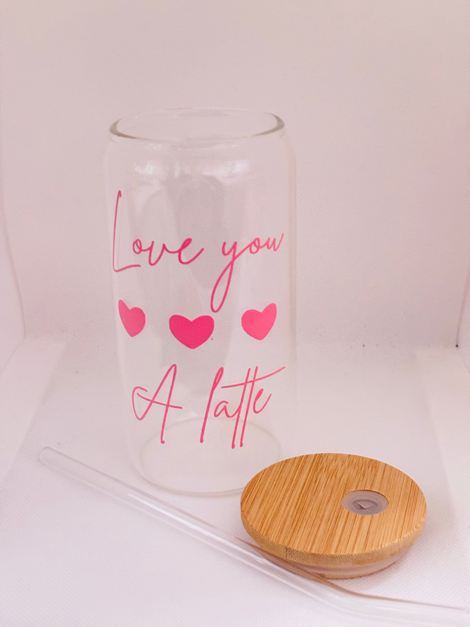 Clear glass with pink text 'Love you a latte' and hearts, next to a wooden lid and straw on a white background.
