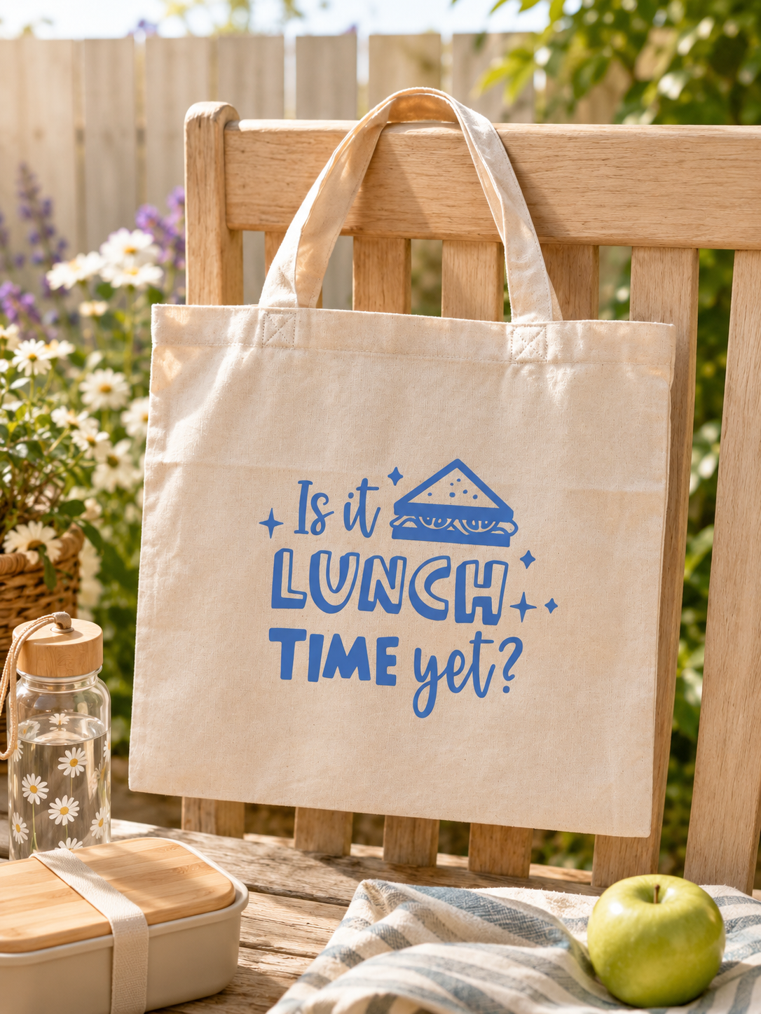 Tote bag with 'Is it lunch time yet?' text on a wooden chair outdoors.