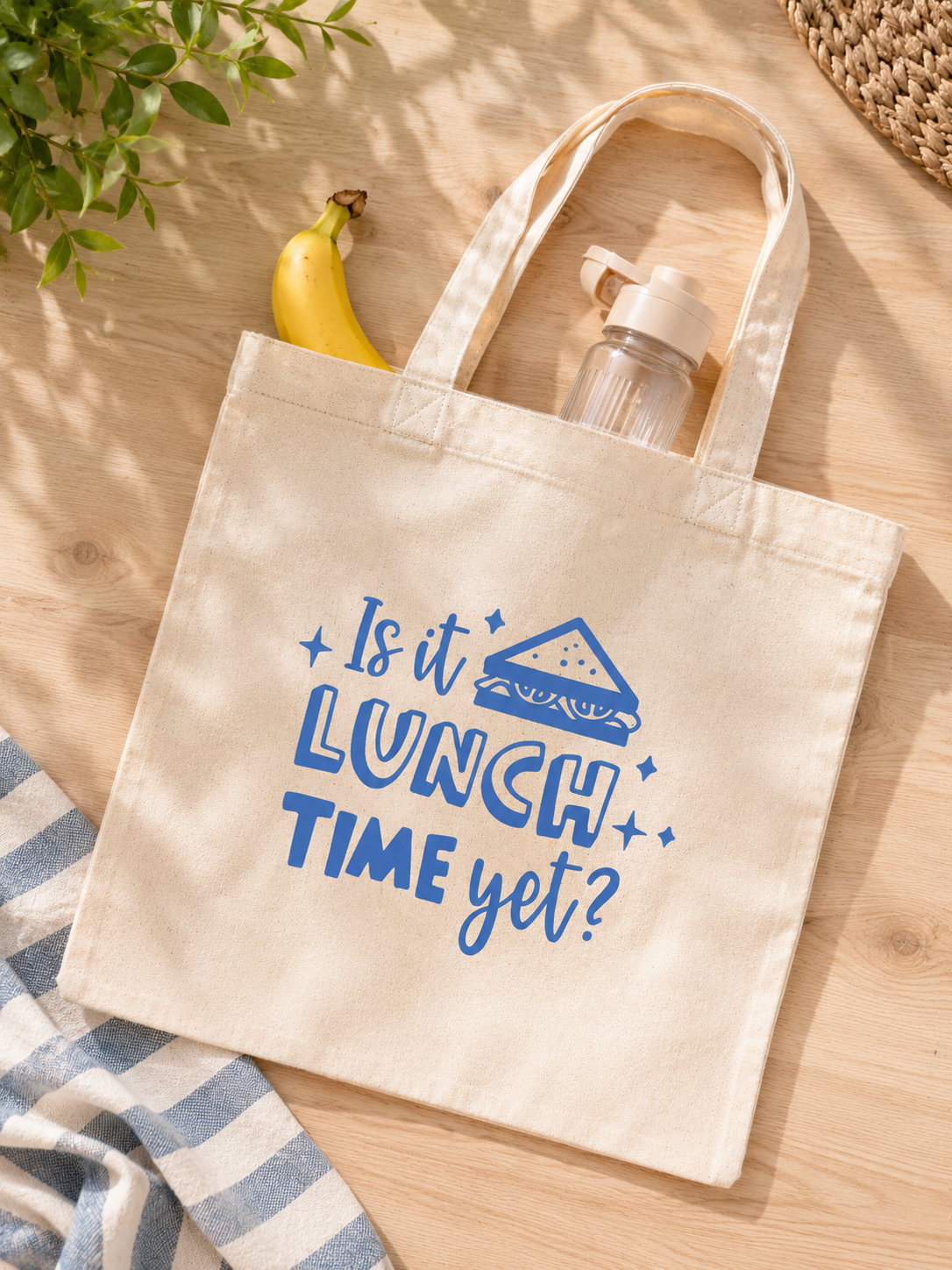 Beige tote bag with 'Is it lunch time yet?' text on a wooden surface with a banana and water bottle.