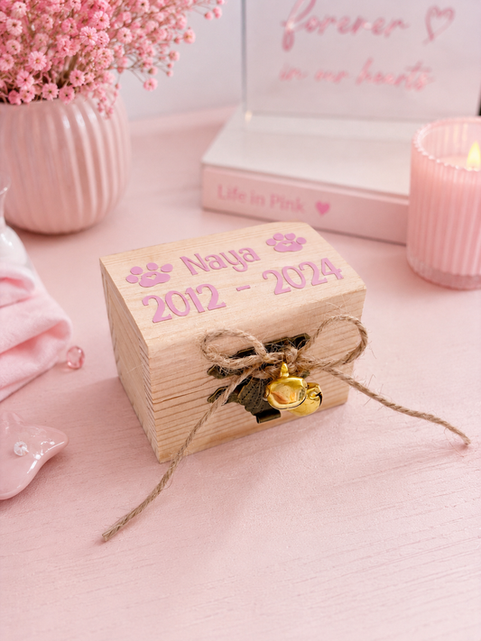 A small personalized wooden keepsake box with 'Naya 2012 - 2024' and paw prints on the top, tied with a twine bow and a small gold bell, set on a pink tabletop with pink flowers and candles in the background.