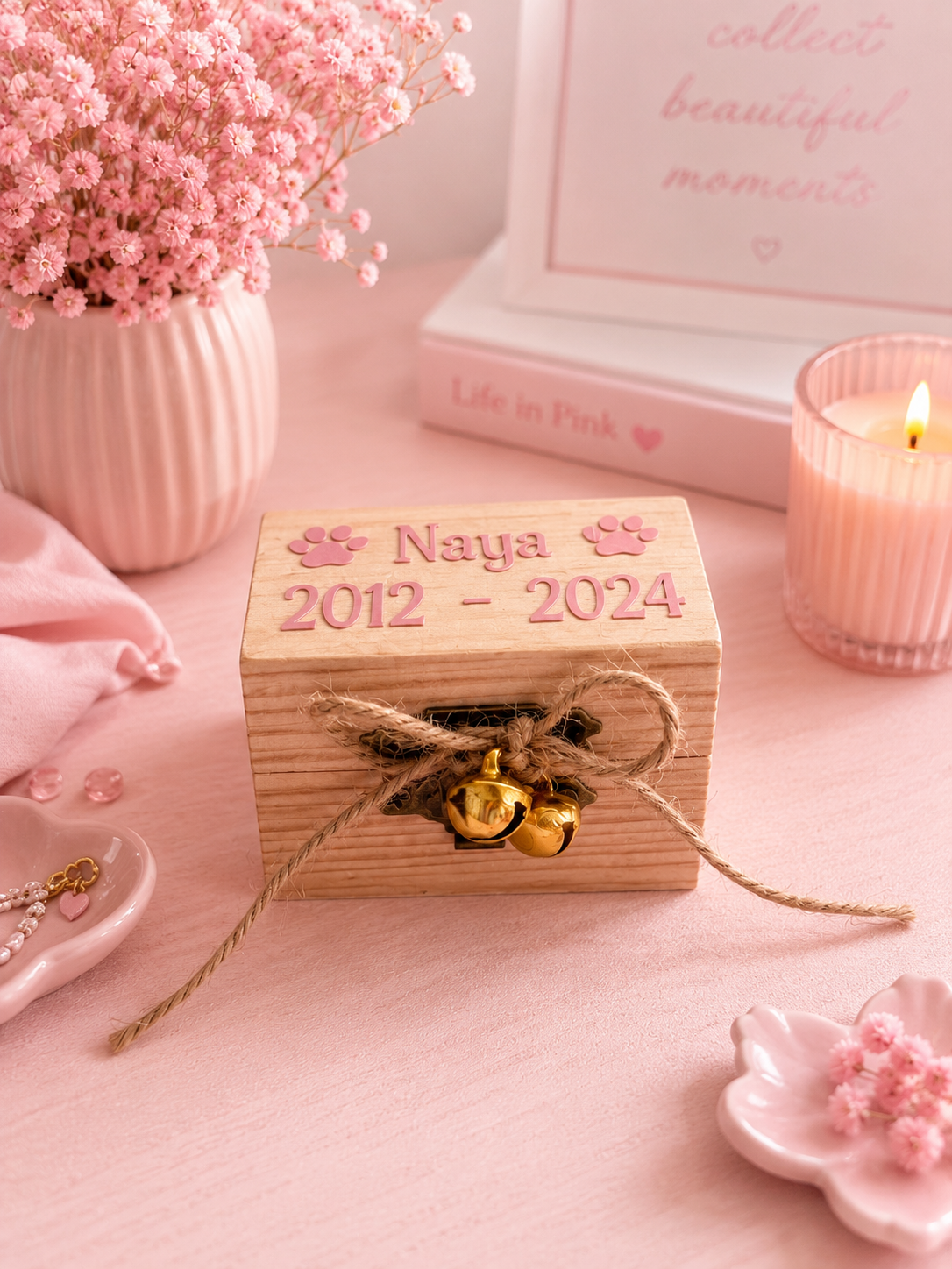 A small wooden keepsake box with 'Naya 2012 - 2024' and paw prints, tied with twine and gold jingle bells, styled on a pink surface with flowers, a candle, and books in the background.