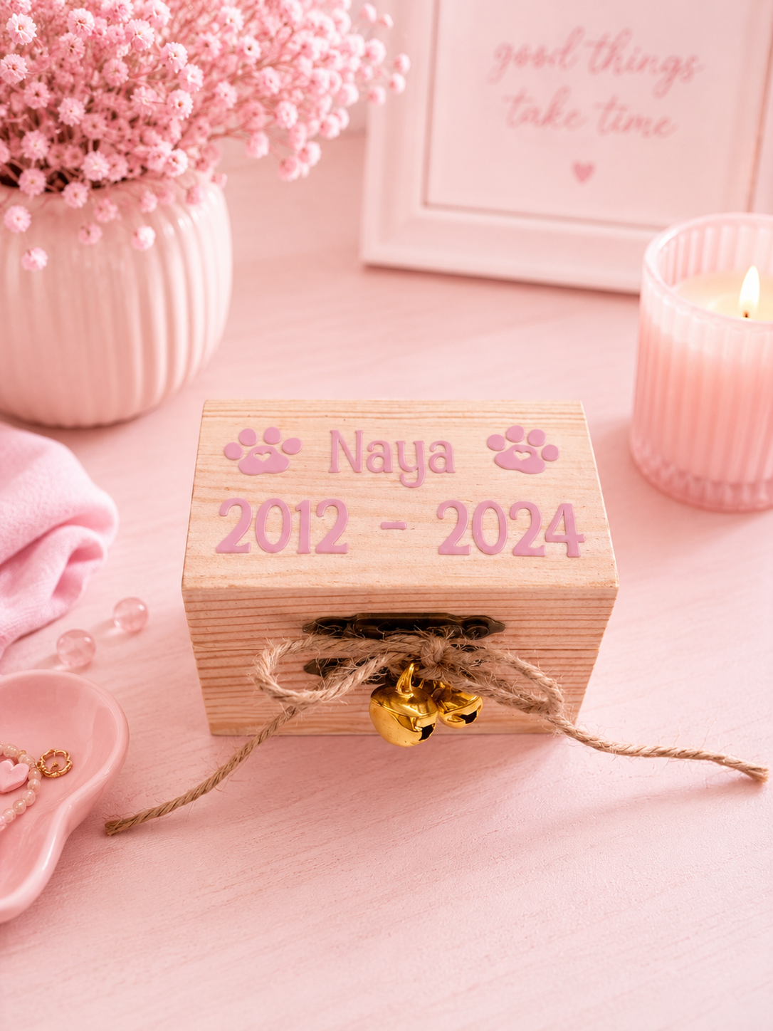 A small wooden keepsake box with 'Naya 2012 - 2024' and paw prints, tied with twine and a small gold bell, styled on a pink surface with flowers, a candle, and a framed print in the background.