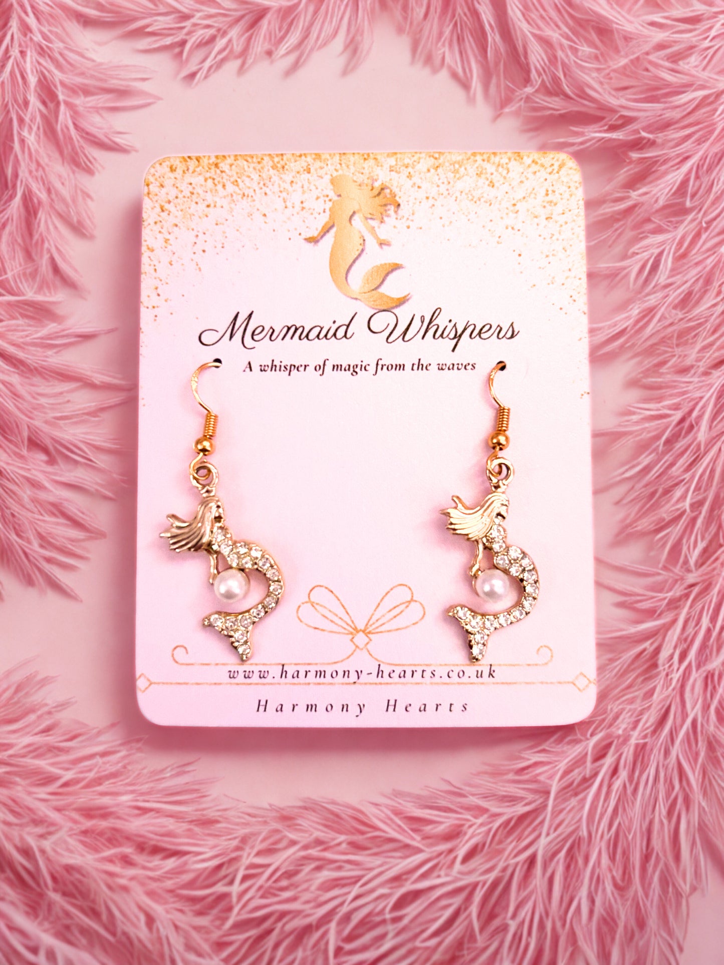 Gold mermaid-shaped earrings on a 'Mermaid Whispers' branded card against a pink fluffy background.