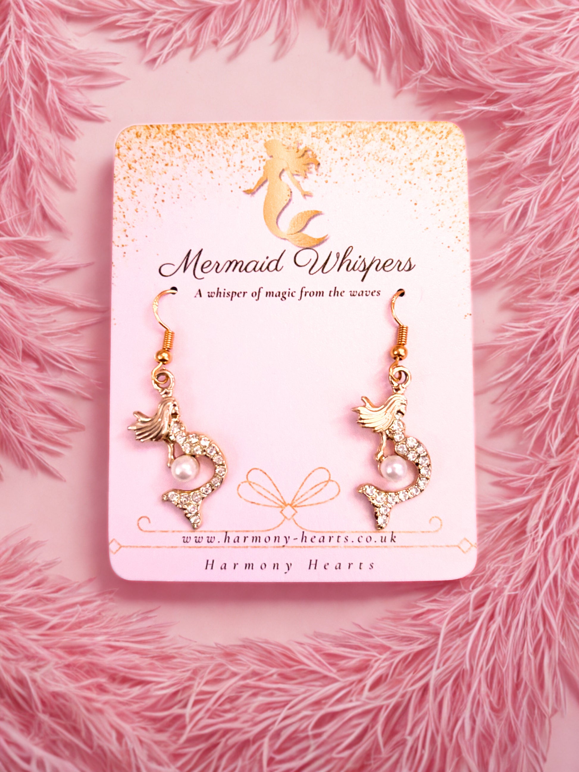 Gold mermaid-shaped earrings on a 'Mermaid Whispers' branded card against a pink fluffy background.