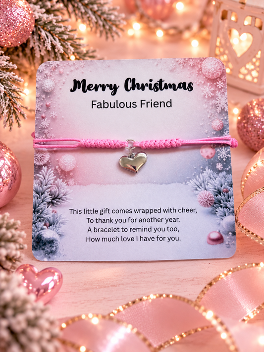 A pink knotted cord bracelet with a small silver heart charm, presented on a printed holiday gift card that reads 'Merry Christmas Fabulous Friend.' The card is styled with pink ribbons, glitter ornaments, and warm string lights in a festive setting.