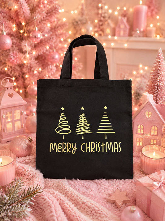 A black canvas tote bag with gold 'Merry Christmas' text and three Christmas tree graphics, displayed in a cozy pink holiday setting with candles, lights, and gift boxes.