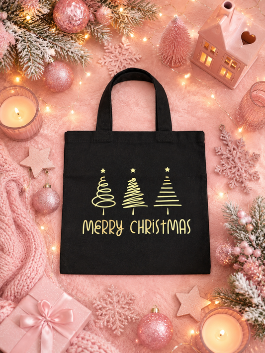 A black canvas tote bag with gold 'Merry Christmas' text and three gold Christmas tree graphics, displayed in a festive flat-lay setting with pink holiday decorations, candles, and a small house ornament.