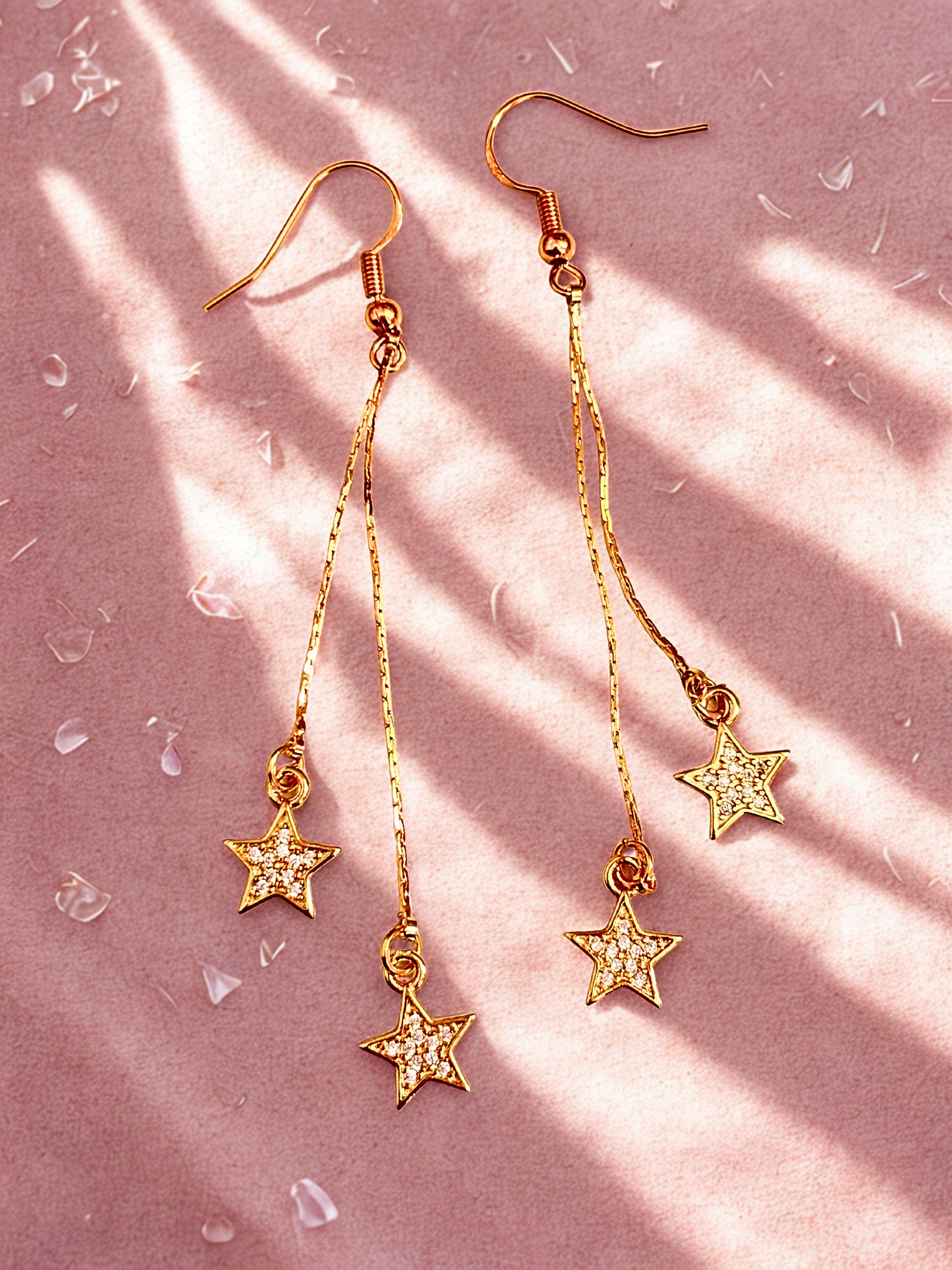 Gold drop earrings with sparkly stars laid on a pink surface