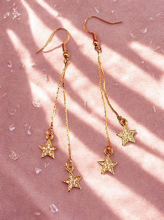 Gold drop earrings with sparkly stars laid on a pink surface