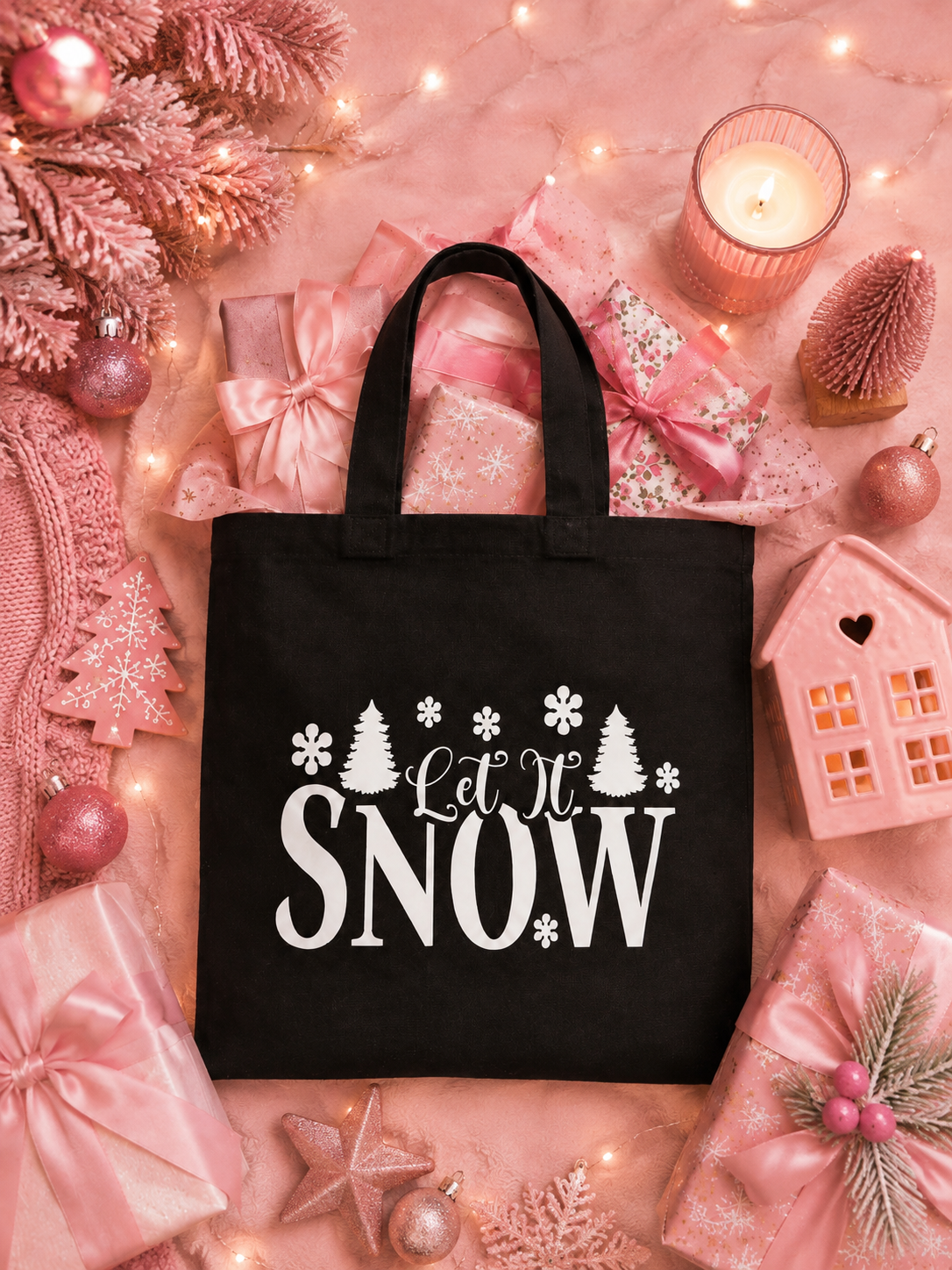 A black canvas tote bag with white 'Let It SNOW' text and holiday graphics, styled in a pink Christmas flat lay with wrapped gifts, ornaments, a candle, and decorative lights.