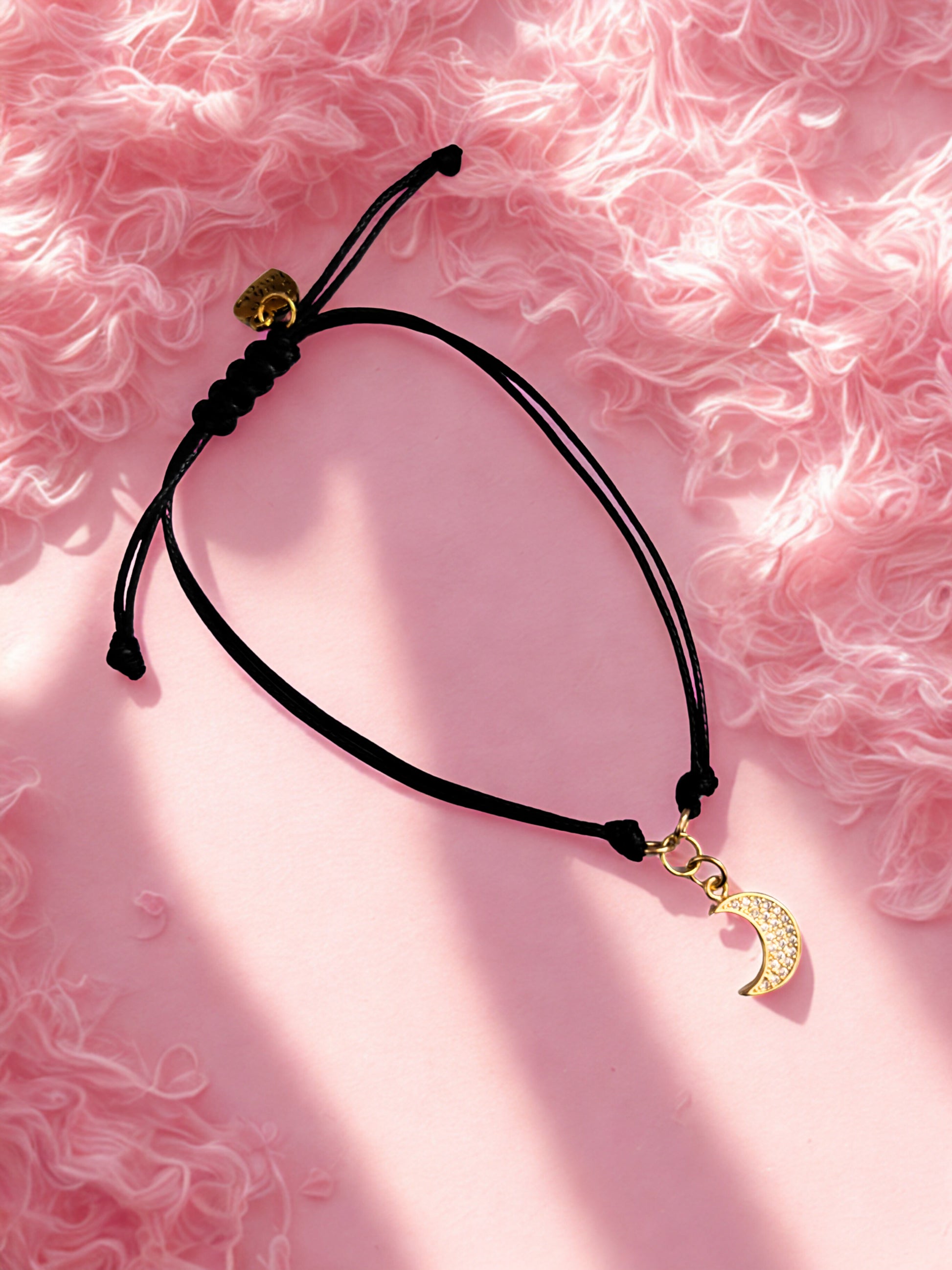 Black cord bracelet with gold crescent moon charm on a pink textured background