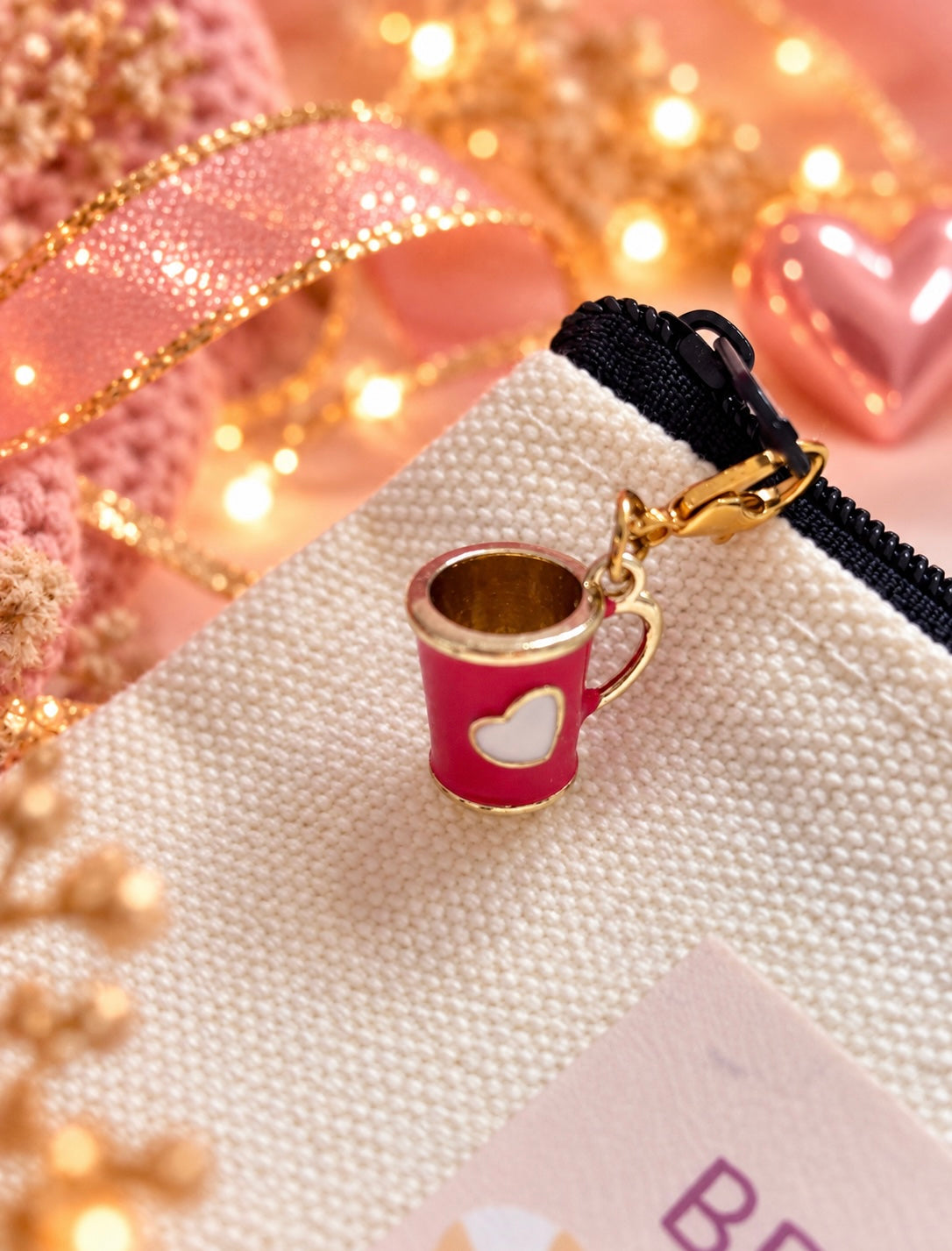 A small enamel charm shaped like a pink coffee cup with a white heart and gold-tone trim, attached to a black-and-white zipper pull on a cream woven pouch. The scene is styled with pink glitter ribbon, warm fairy lights, and pink decorative elements in the background.