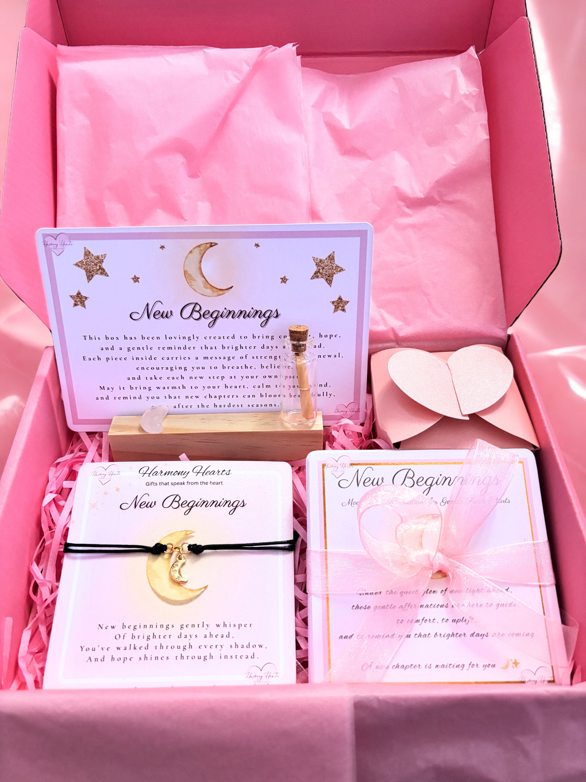 Gift set with 'New Beginnings' card, bracelet, and heart-shaped object in a pink box.