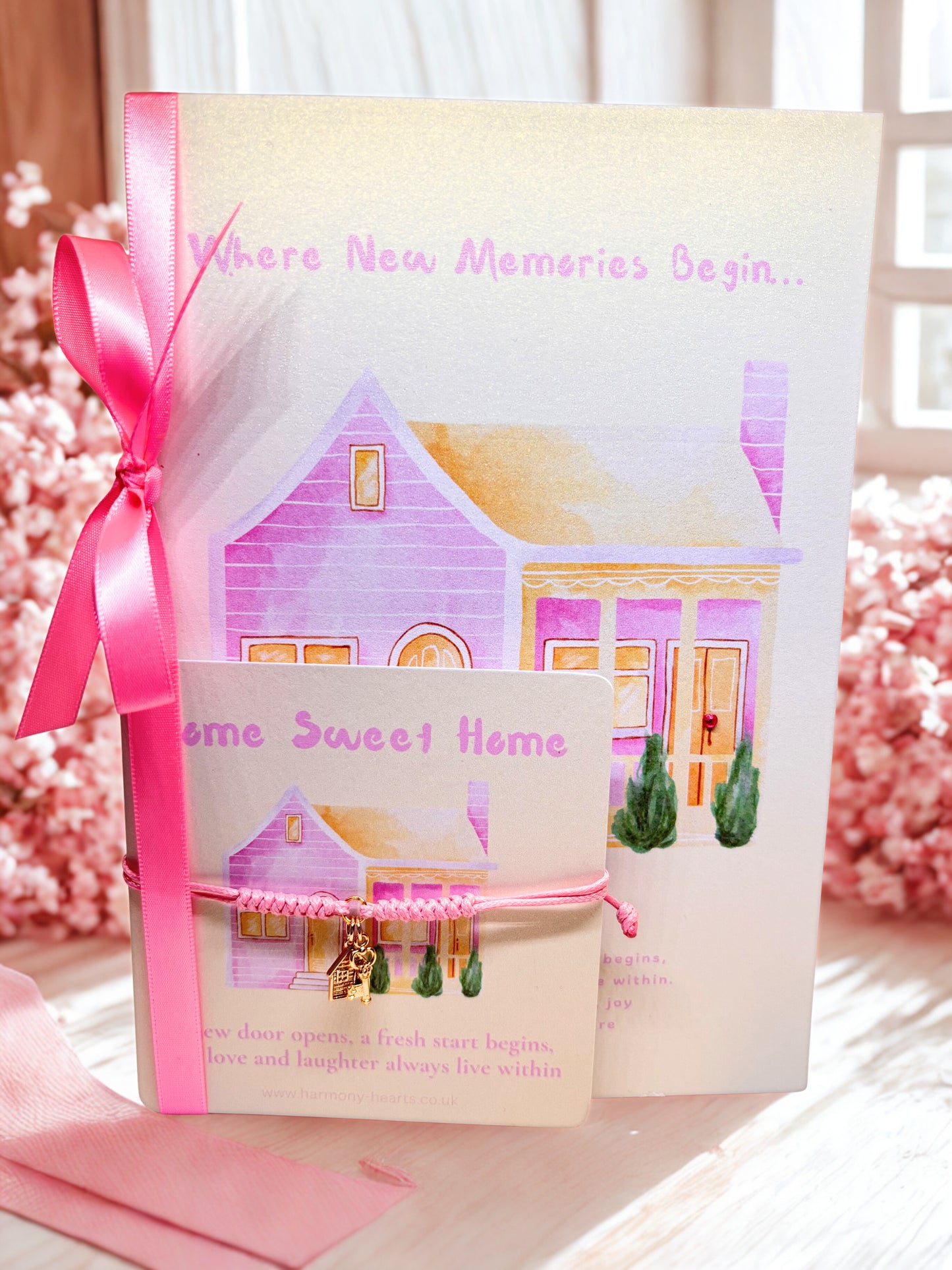 Gift card with house illustration and pink ribbon and a pink cord charm bracelet on a matching card on a decorative background