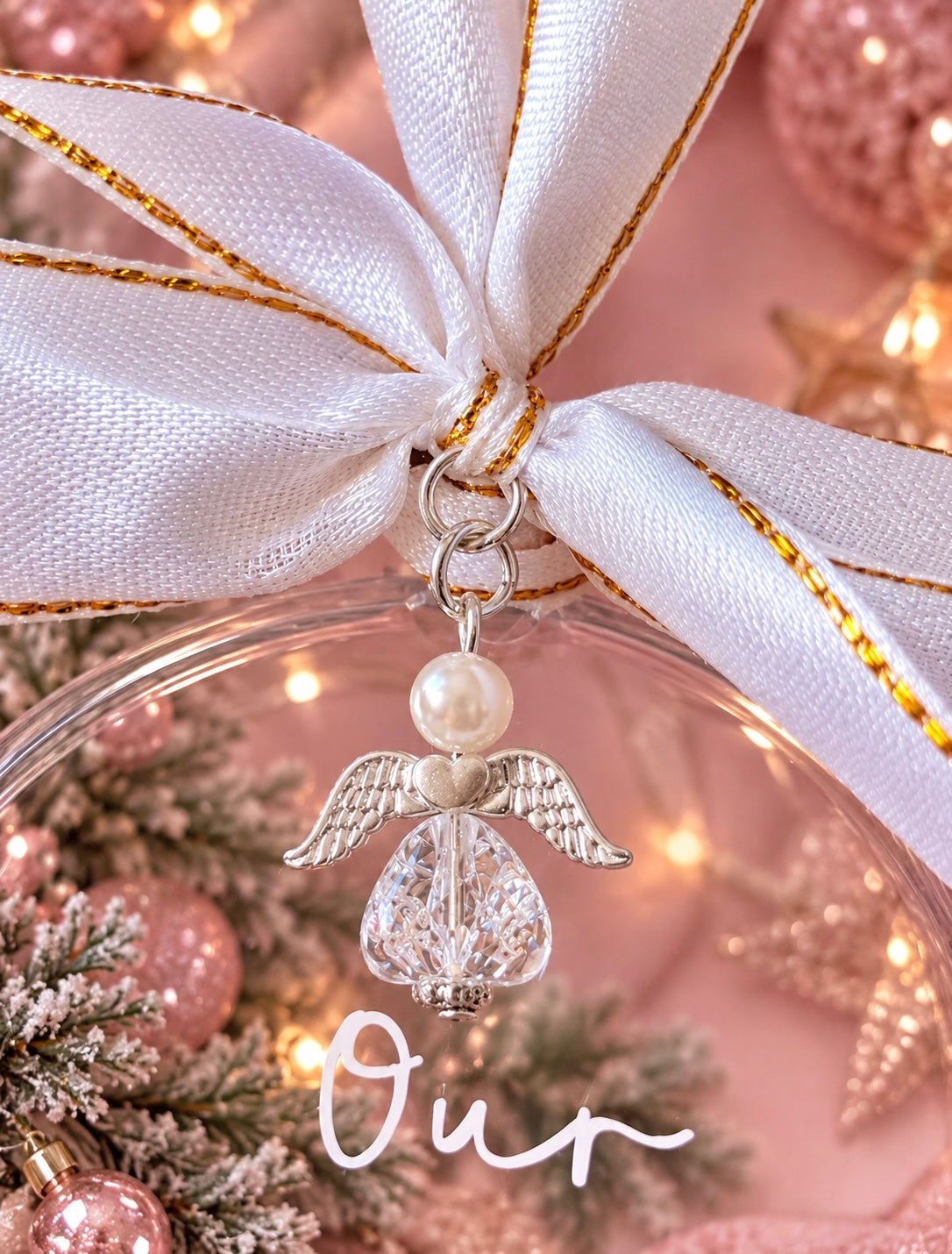 A close-up of a clear round ornament tied with a large white satin ribbon with gold trim. Hanging from the ribbon is a silver angel charm with a pearl-like head and a faceted clear body.