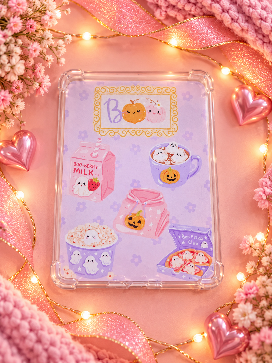 A clear protective kindle case with a cute pastel Halloween-themed insert, displayed on a pink background decorated with pink ribbons, fairy lights, and small flowers.