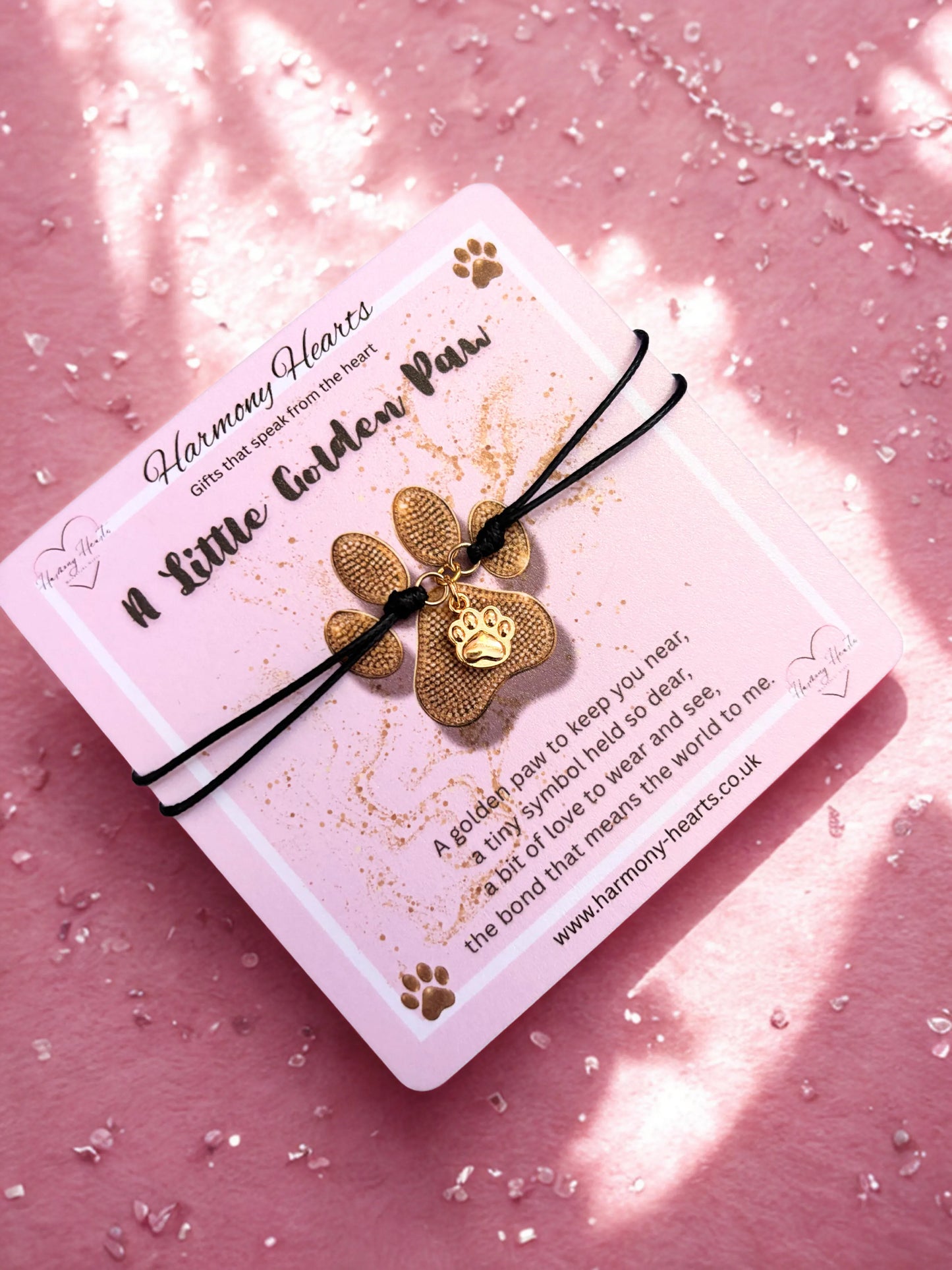 Gold paw-shaped charm bracelet on a black cord with a card on a pink background