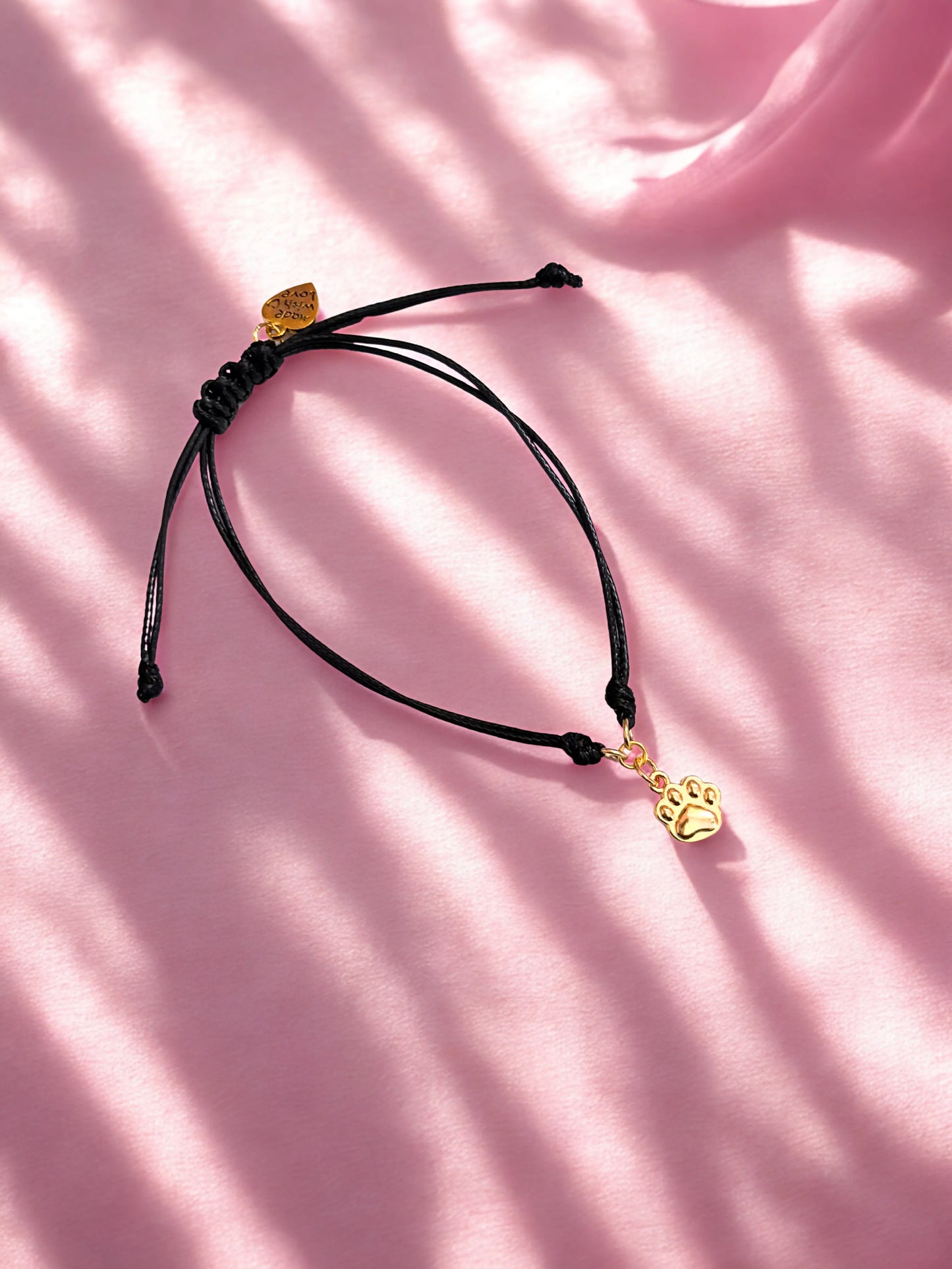 Black cord bracelet with a gold paw charm on a pink background