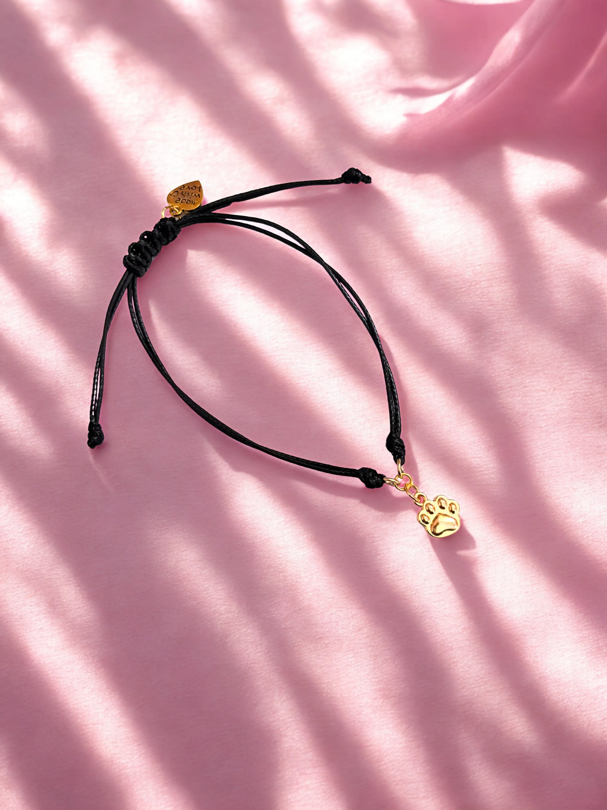 Black cord bracelet with a gold paw charm on a pink background