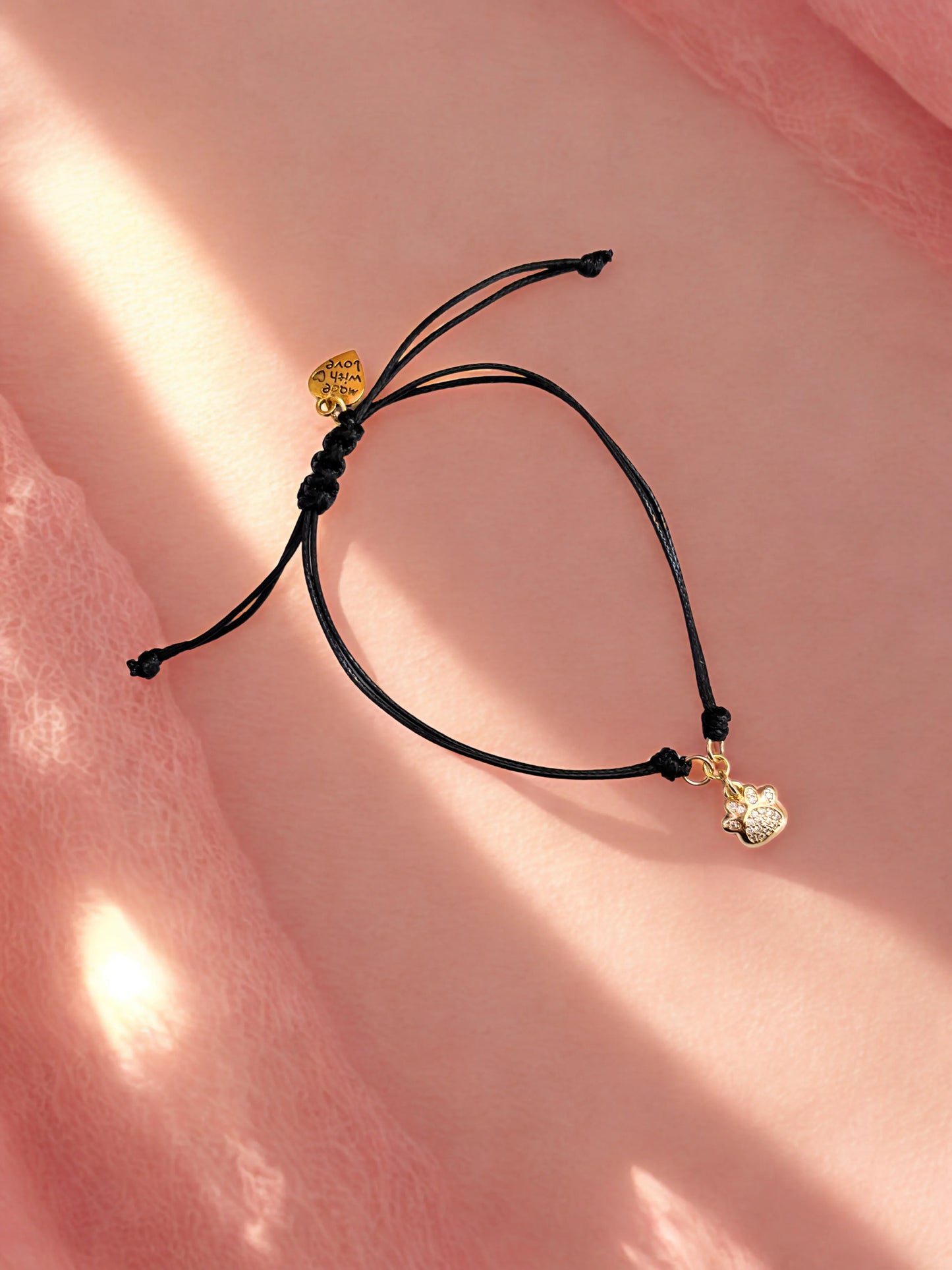 Black cord bracelet with gold paw charm on a pink background