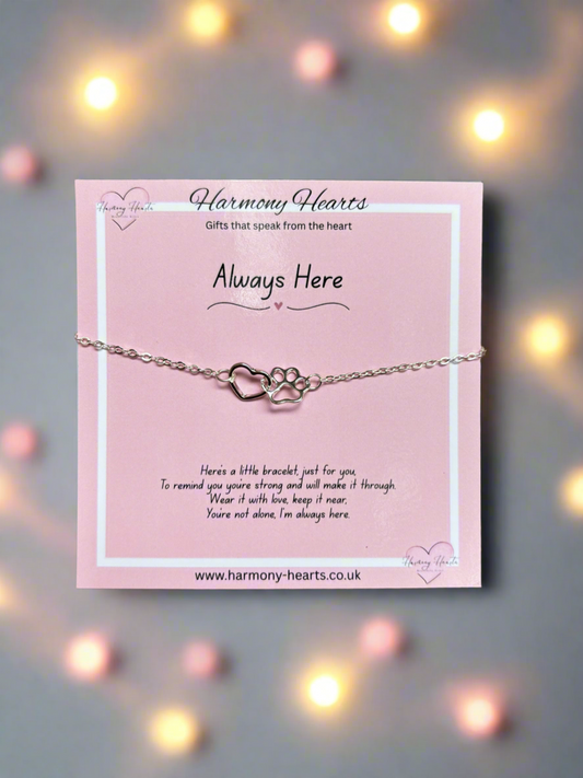 Silver bracelet with heart and paw design on a pink card with 'Harmony Hearts' branding, set against a blurred background with lights.