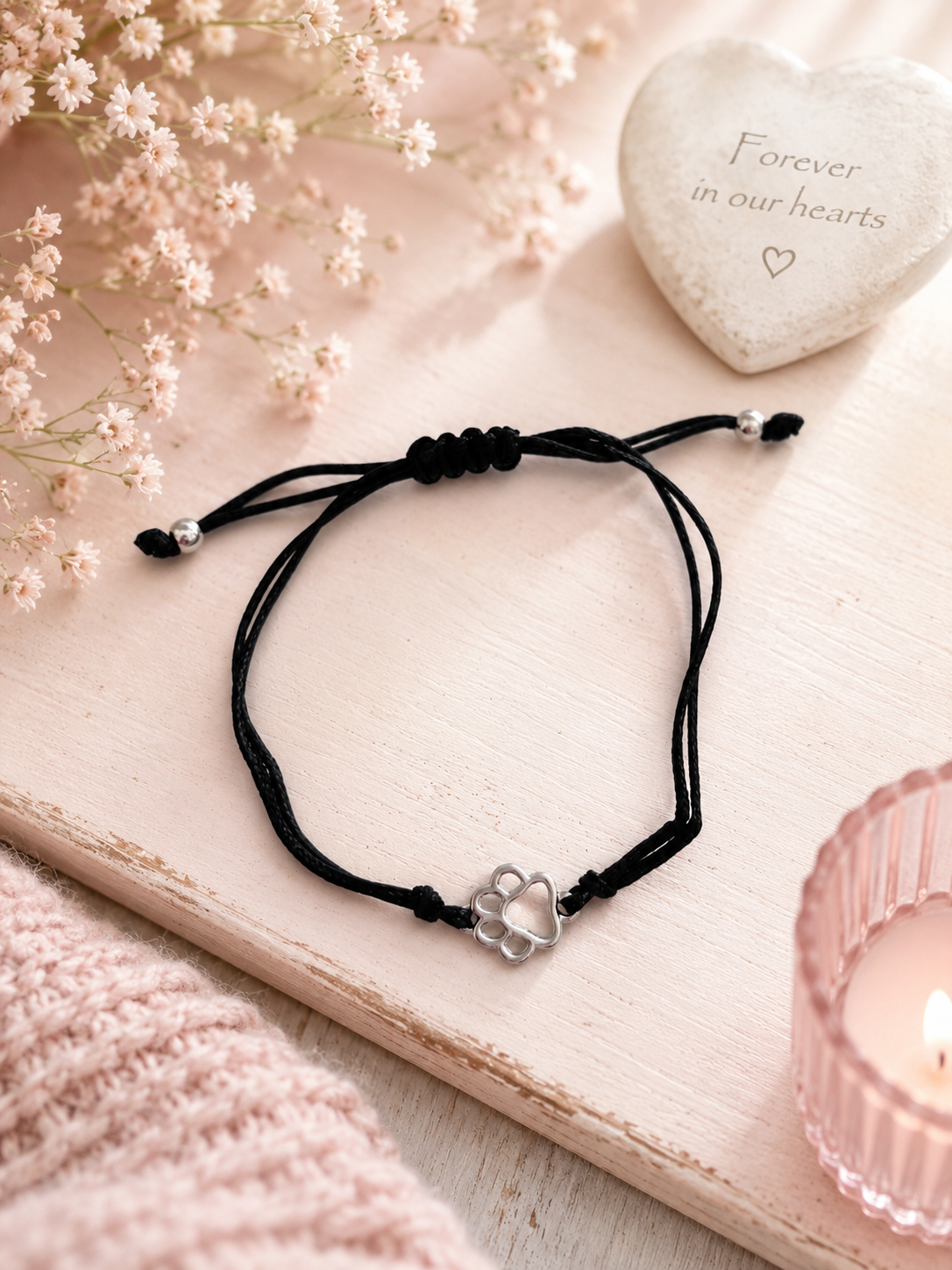 A delicate black cord bracelet with a silver paw print charm, styled on a light pink wooden board with dried flowers, a heart-shaped stone, and a pink candle.
