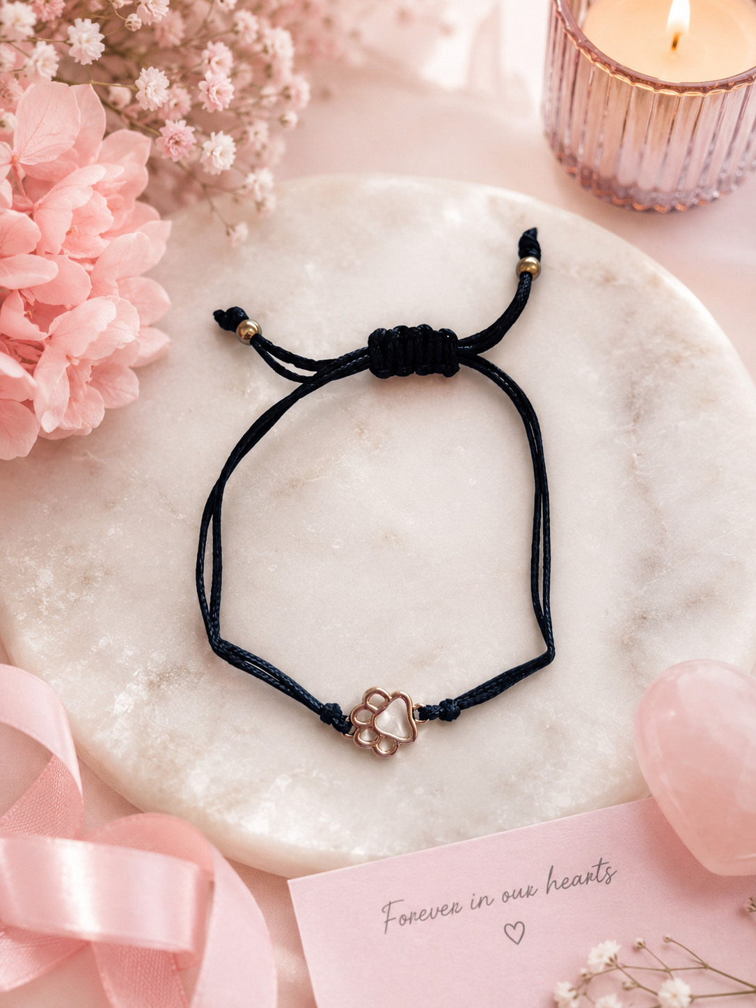 A black cord adjustable bracelet with a small paw print charm, displayed on a round white marble tray surrounded by pink flowers, a lit candle, and a pink card reading 'Forever in our hearts'.