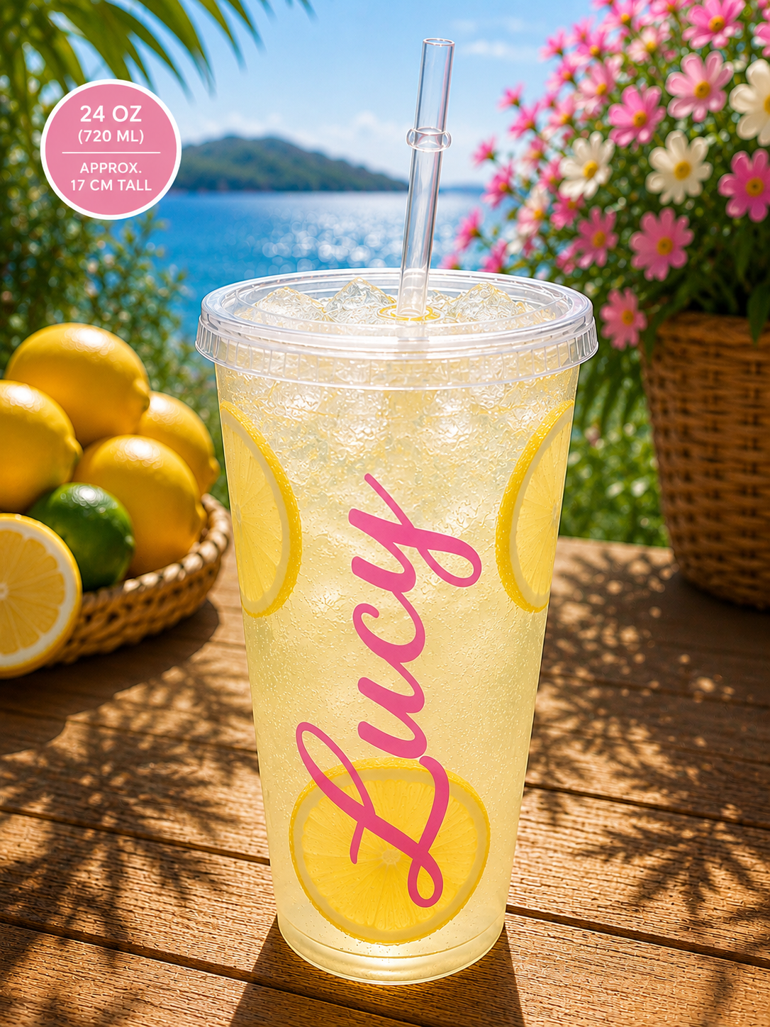 A clear 24 oz plastic travel tumbler cup with a clear straw, filled with a lemonade drink and decorated with yellow lemon slices and a pink script name 'Lucy'. The cup is placed on a wooden table outdoors with lemons, flowers, and a beach background.