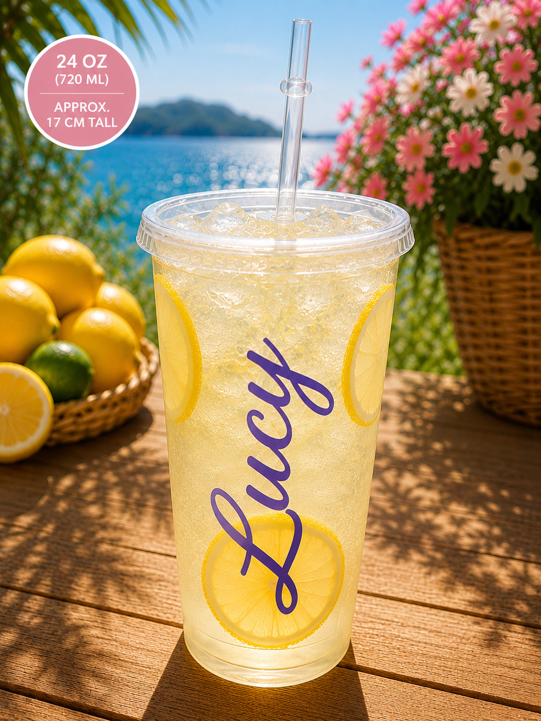 A clear 24 oz plastic tumbler cup with a straw, filled with a lemon drink and decorated with a purple script name reading 'Lucy'. The cup is placed on a wooden table outdoors with lemons, flowers, and a body of water in the background.